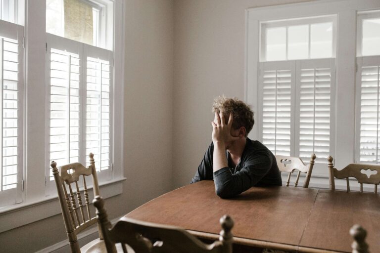 Man sitting at a kitchen table with his hand on his face while looking out a window, reflecting doubt and rumination often explored in therapy for ROCD in Denver, CO.