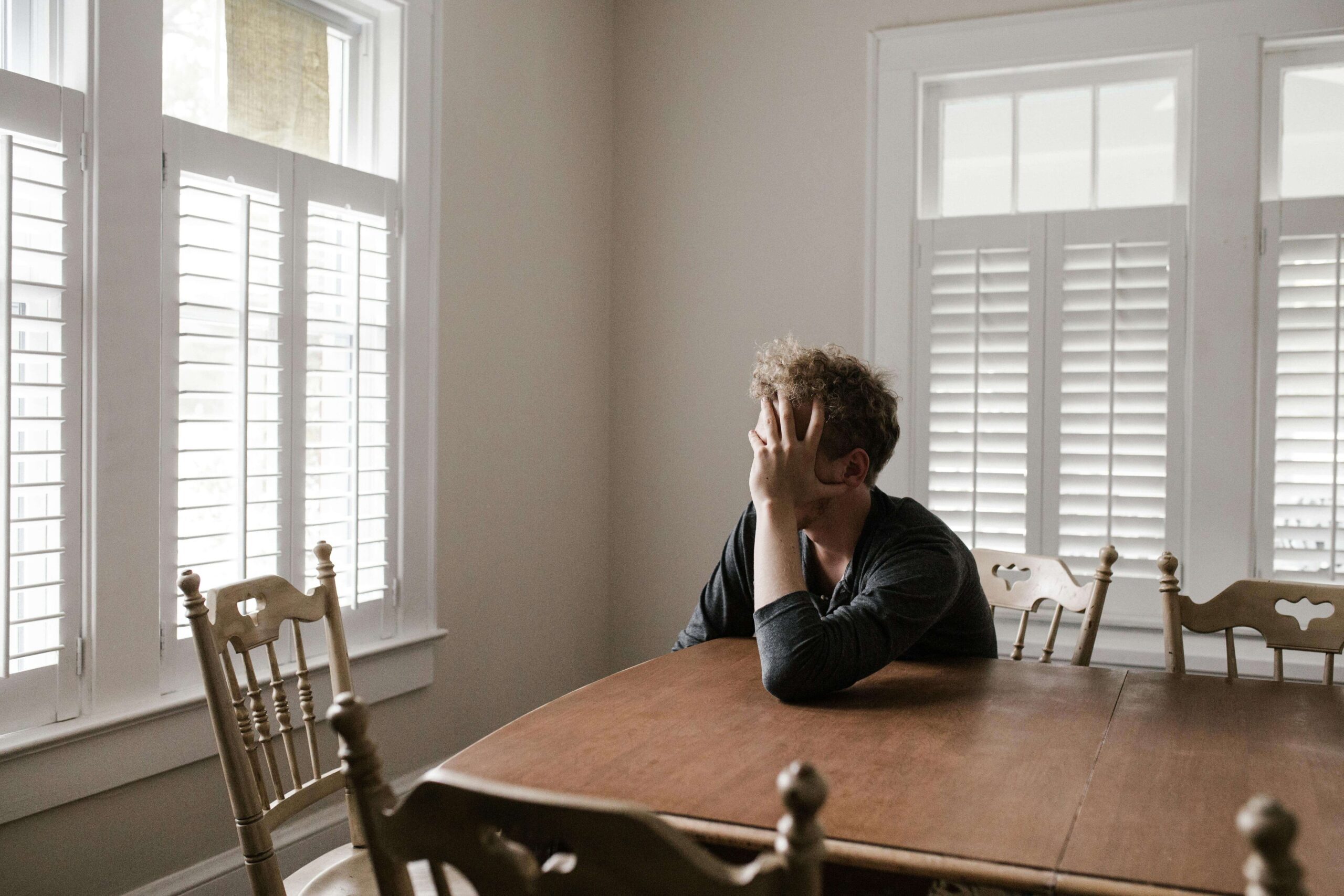 Man sitting at a kitchen table with his hand on his face while looking out a window, reflecting doubt and rumination often explored in therapy for ROCD in Denver, CO.