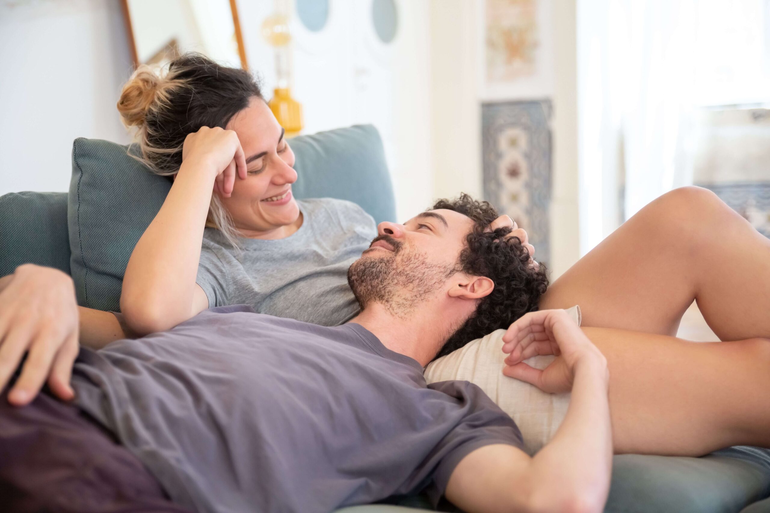Couple lying on a couch together with the man resting on the woman’s lap while they look at each other happily, symbolizing trust and closeness fostered through individual relationship counseling for emotional security in Denver, CO.